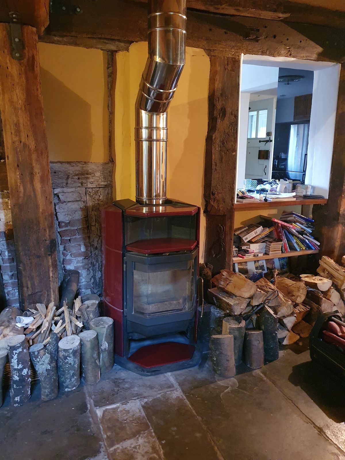 A stove with a stainless flue in a timber framed room beside a log store